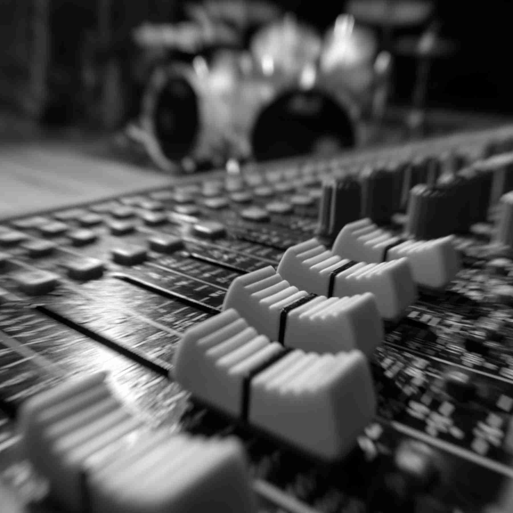 Black and white photograph of mixing board faders in the foreground with an out of focus drum kit in the background.